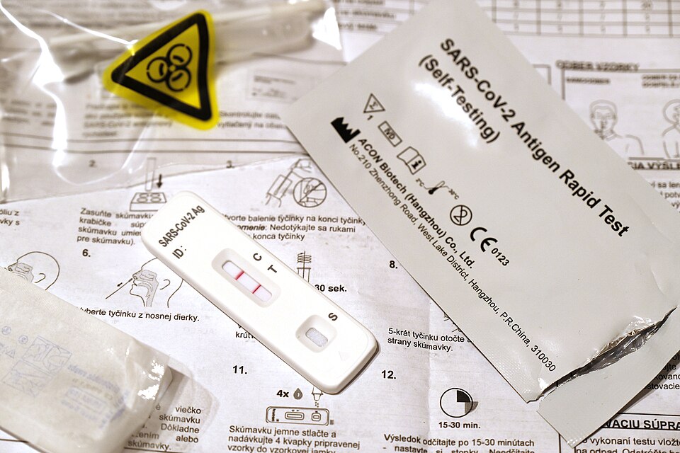 Multiple items are arranged on a surface covered with printed instructions. A test cassette displays two pink lines next to letters "C" and "T" along with "SARS-CoV-2 Ag" printed on it, indicating a COVID-19 self-test. Next to it, a torn foil packet reads "SARS-CoV-2 Antigen Rapid Test (Self Testing)" with manufacturer details: "ACON Biotech (Hangzhou) Co., Ltd" and other symbols. A plastic bag contains a biohazard symbol—a black circle with three circular shapes inside, encircled by yellow. An unopened pad or wipe packet is partially visible. The background features diagrams and text, possibly in a Slavic language, providing instructions for the test.