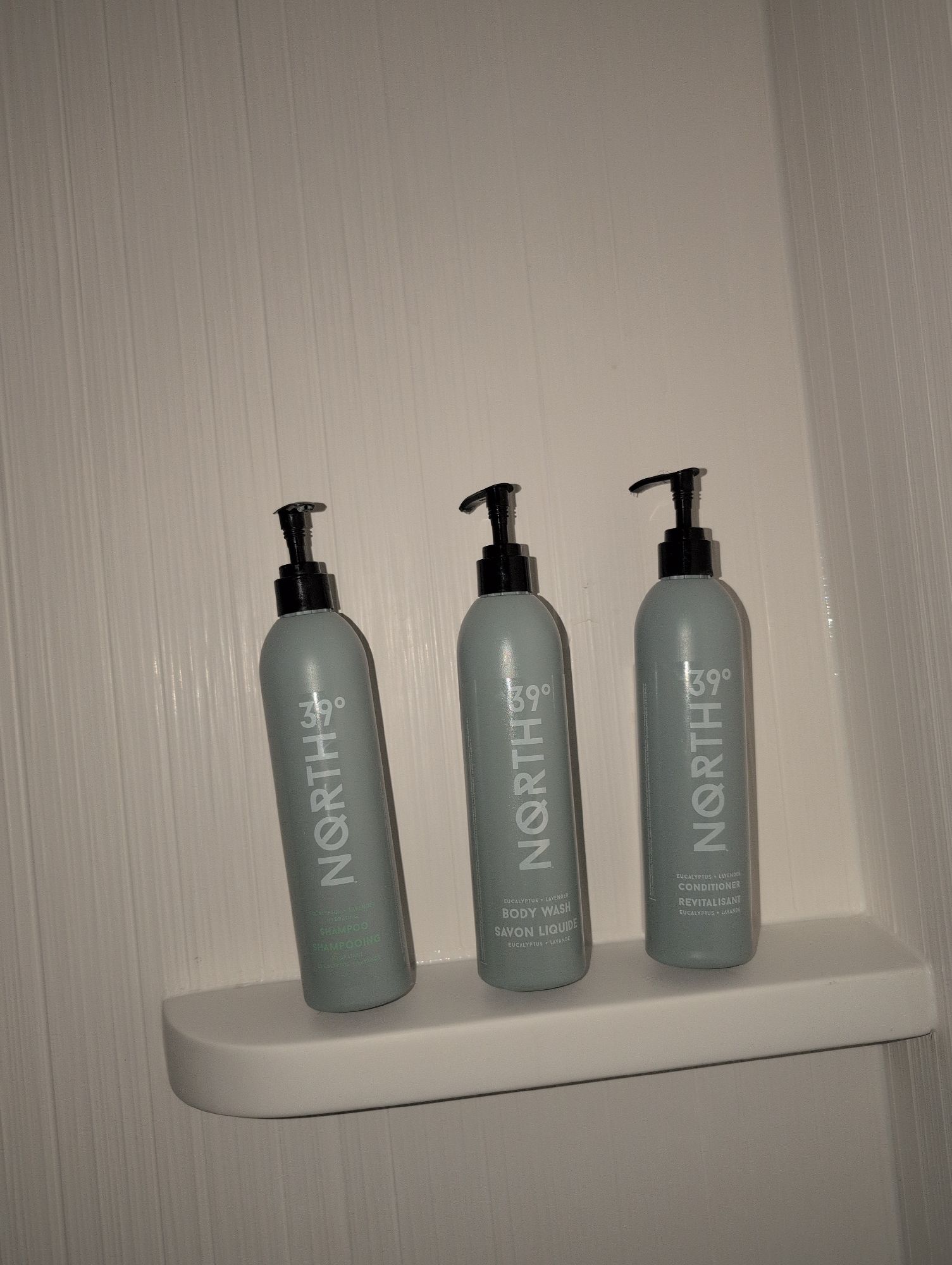 A shower corner with three matching light gray-green pump bottles arranged side by side on a small white corner shelf against vertically ridged white shower walls; the photo appears to be taken with flash, creating bright reflections on the bottles and deep shadows behind them. Each bottle has a black pump and the brand text “NORTH 39°” printed vertically in white. From left to right, the labels read: “SHAMPOO / SHAMPOOING,” “BODY WASH / SAVON LIQUIDE,” and “CONDITIONER / REVITALISANT,” with smaller text “EUCALYPTUS + LAVENDER” near the bottom of the body wash and conditioner. The composition is simple and centered on the three bottles, with no other objects visible.