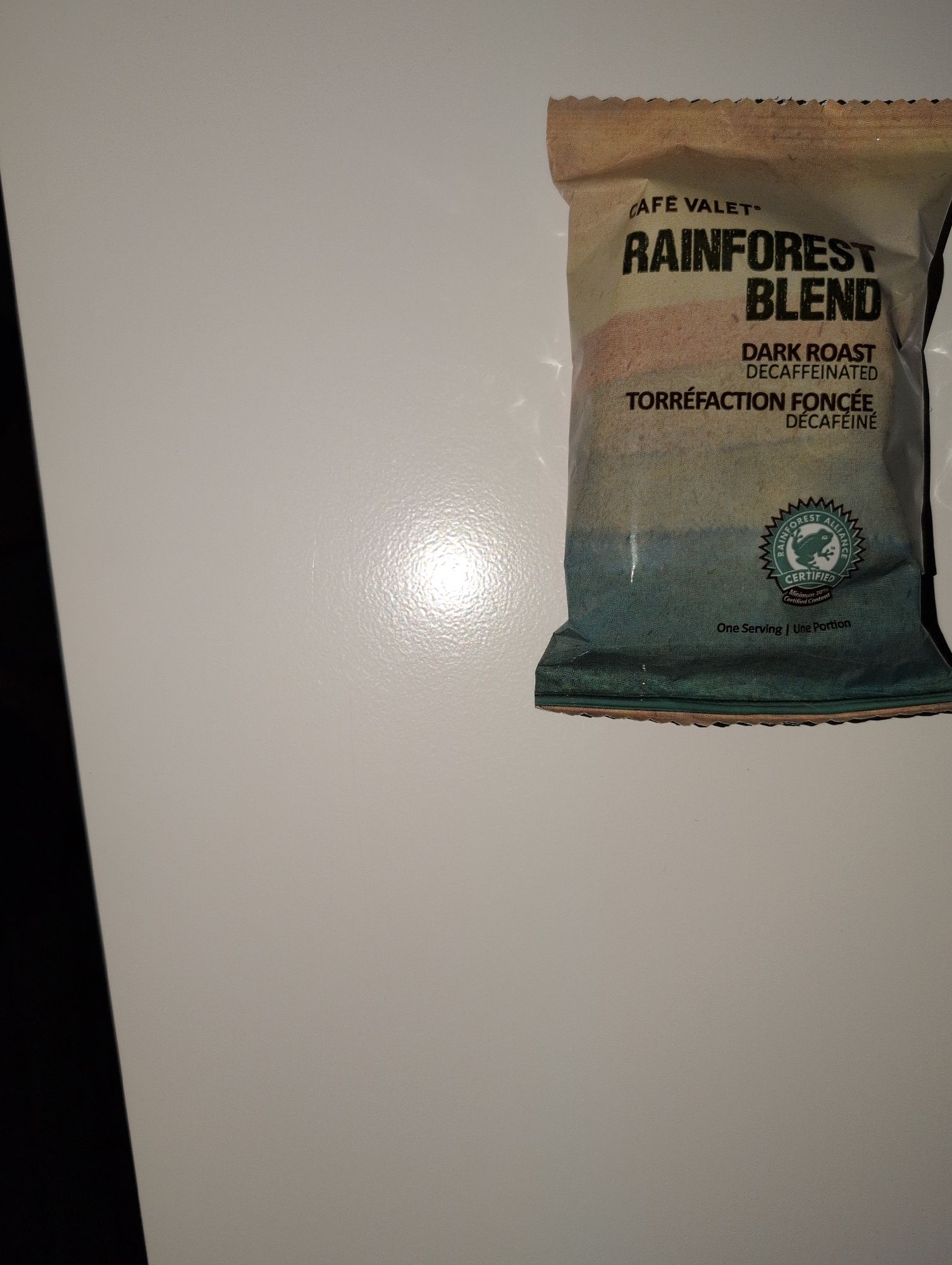 The right half of a small, single‑serve coffee packet lying on a glossy white surface, with most of the frame taken up by the empty surface and a bright circular glare from the camera flash near the center-left; the background beyond the surface is dark. Only the right portion of the packet is visible at the upper right edge, with a tan‑to‑teal gradient design and crimped top and bottom edges. Legible text on the packet reads: “CAFE VALET,” “RAINFOREST BLEND,” “DARK ROAST DECAFFEINATED,” “TORRÉFACTION FONCÉE DÉCAFÉINÉ,” and “One Serving | Une Portion.” A green circular seal shows the words “RAINFOREST ALLIANCE CERTIFIED” around a frog logo. The image is sharply focused with harsh, direct lighting from the flash and is taken from a slightly elevated angle.