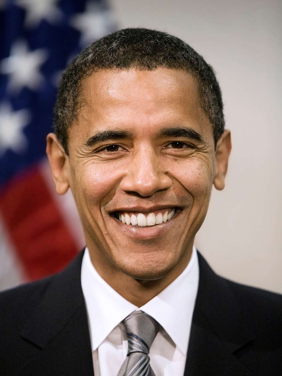 A well-lit, close-up portrait shows President Barack Obama, an adult man facing the camera from the chest up, centered in the frame, with a shallow depth of field that blurs the background. He has short, closely cropped dark hair, dark eyes, and a smooth-shaven face. He is smiling with teeth visible, and faint smile lines are visible around his eyes and mouth. He wears a dark suit jacket, a white dress shirt, and a gray striped tie. Behind him, out of focus, is part of a United States flag with white stars on a blue field and red-and-white stripes. The image is sharp and evenly exposed, photographed straight on at eye level, with no visible text.