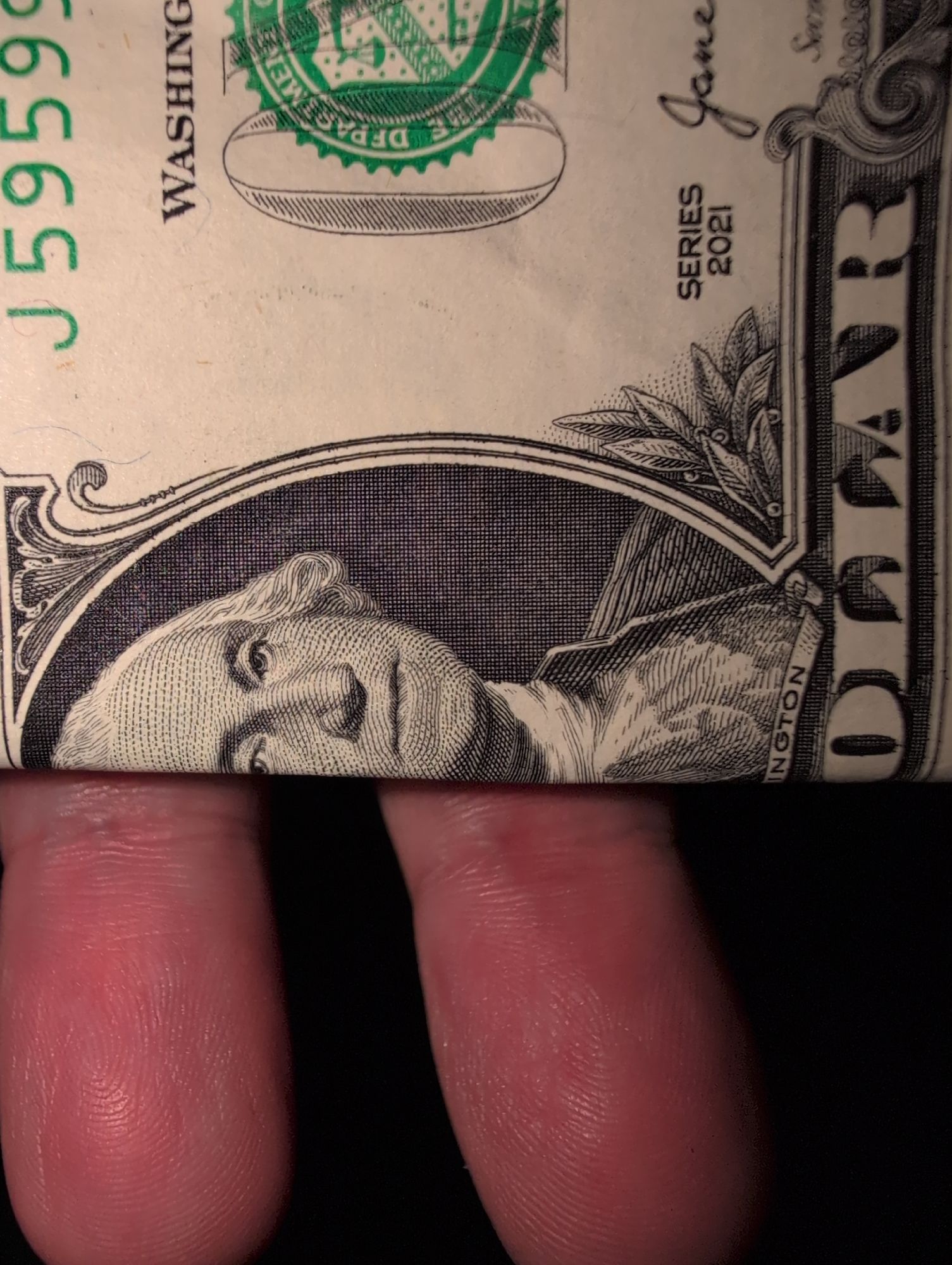 Close-up of a folded U.S. one-dollar bill being held between two fingers. The bill shows part of the portrait of George Washington, the green Treasury seal, and part of the serial number. The texture and detail of the skin on the fingers holding the bill are also clearly visible. The bill is from the 2021 series.