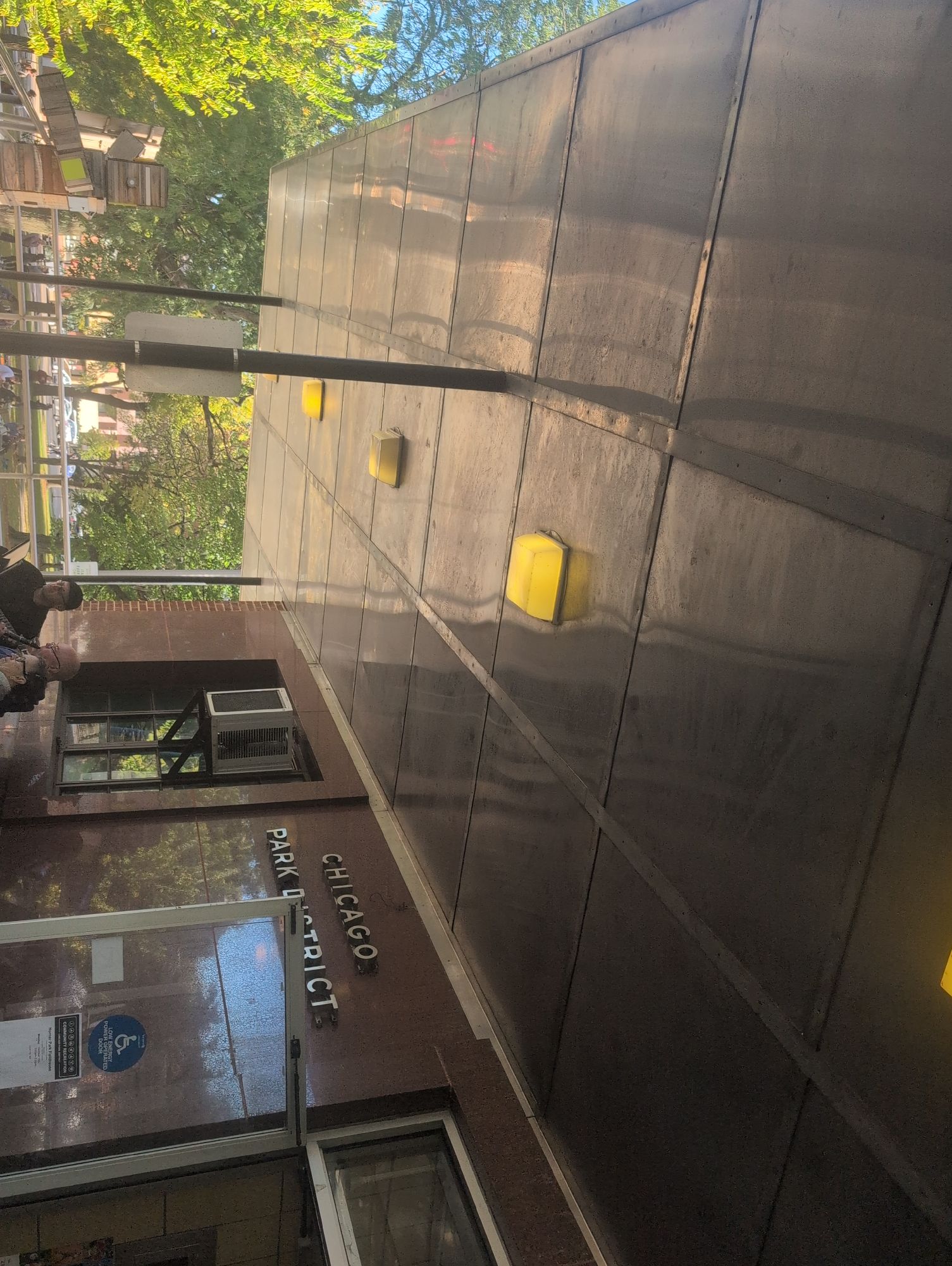 A sidewalk slopes upward with a large wall on the right featuring yellow rectangular lights at regular intervals. On the left, a brick building has signage that reads "CHICAGO PARK DISTRICT." Below this sign is a window with an air conditioning unit. Near the bottom, a person wearing a dark-colored jacket and carrying a backpack is visible, standing close to the building. Green trees with sunlight filtering through are in the background, casting dappled light on the scene.