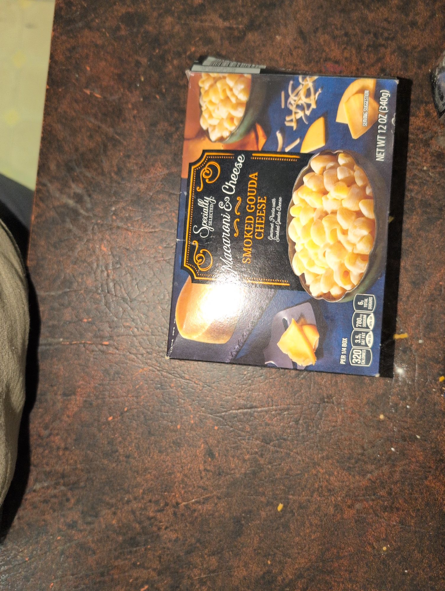 Box of macaroni and cheese on a brown surface. The packaging reads "Specially Selected Macaroni & Cheese Smoked Gouda Cheese". The front shows an image of creamy macaroni in a bowl, along with slices and shreds of smoked gouda cheese. Nutritional information is visible in the bottom right corner, indicating 320 calories per serving. The net weight of the package is 12 oz (340g).