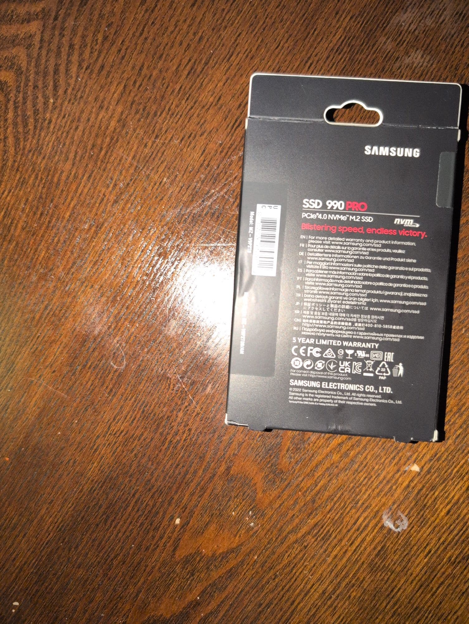 The back of a black retail box for a Samsung solid-state drive (SSD), resting on a wooden surface. The surface has a prominent, dark brown wood grain pattern running diagonally and shows some minor signs of wear and a few small, white specks.
The packaging itself is for a Samsung SSD 990 PRO. This is stated in bold white text, followed by its specifications: "PCIe® 4.0 NVMe™ M.2 SSD". Next to this is the NVM Express logo. Below, in red text, is the product slogan: "Blistering speed, endless victory."
A large portion of the box is dedicated to warranty and product information, written in multiple languages (English, French, German, Italian, Spanish, Portuguese, Polish, Turkish, Japanese, Korean, Chinese, and Russian). Each language directs the user to the website `www.samsung.com/ssd`.
Below the multilingual text, the phrase "5 YEAR LIMITED WARRANTY" is clearly visible. This is followed by a row of various certification and compliance logos, including CE, FC, UKCA, and EAC.
On the left side of the box, a white sticker is partially visible, showing a UPC barcode and the model number, which appears to be Model: MZ-V9P2T0.
At the very bottom of the box, the manufacturer is listed as "SAMSUNG ELECTRONICS CO., LTD." with a copyright notice for the year 2022. Made in Vietnam is written in very small text on the box. The box has a built-in plastic hanger tab at the top for retail display. The overall photograph is taken from a slightly elevated angle.